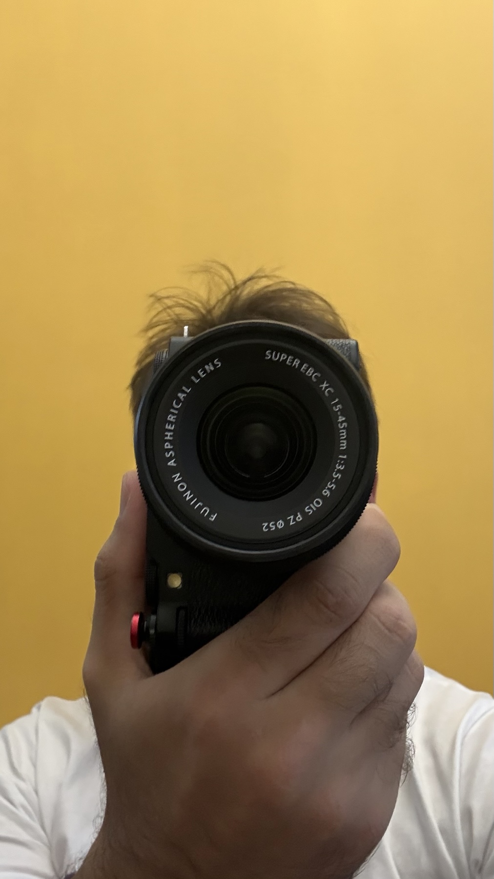 Denis holding a camera in front of his face, against a yellow wall.